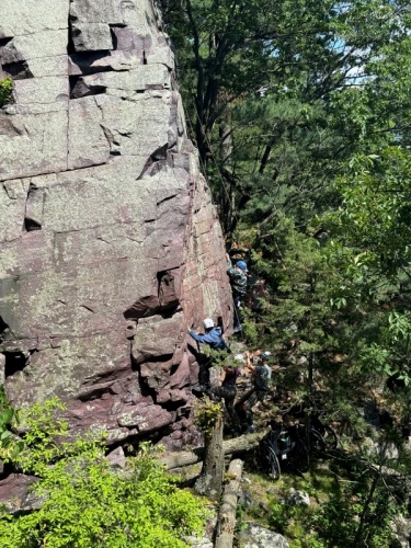 Photo shows two young men wearing climbing helmets and  harnesses part way up a purple quartzite cliff.