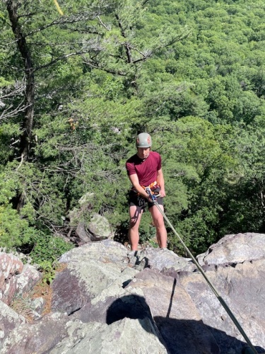 Photo shows a young man wearing a climbing helmet and harness, suspended from ropes in front of a purple quartzite cliff.